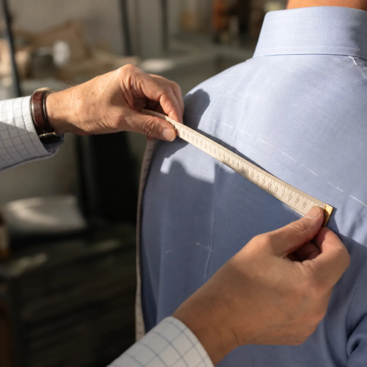 A tailor's hands drawing a tape measure across a client's shoulder in a bright fitting room, brass tape and chalk visible, focus on the cloth and hands.